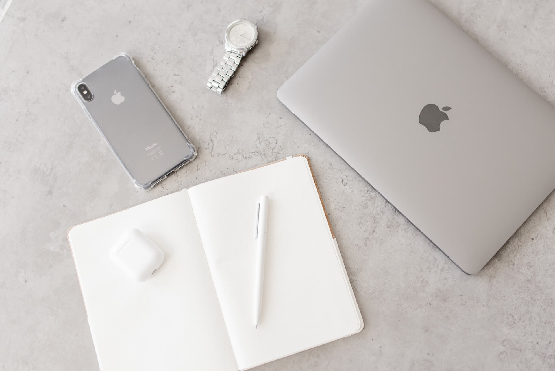 Ein iPhone, eine Armbanduhr, ein MacBook, ein Notizbuch mit einem Stift und kabellose Ohrhörer auf einem Tisch.