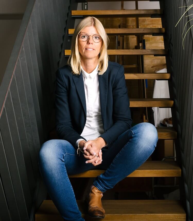 Eine Frau mit blonden Haaren und Brille sitzt auf einer Treppe in einem modernen Büro und blickt in die Kamera.