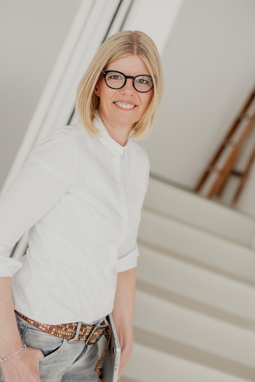 Eine lächelnde Frau mit blonden Haaren und Brille steht in einem modernen Büro. Sie trägt einen dunkelblauen Blazer und Jeans.